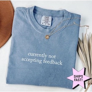 May include: A blue short-sleeved t-shirt with a crew neckline and the white text "currently not accepting feedback" printed on the chest. The shirt features a Comfort Colors brand label at the inner neck and is paired with a gold-toned pendant necklace.