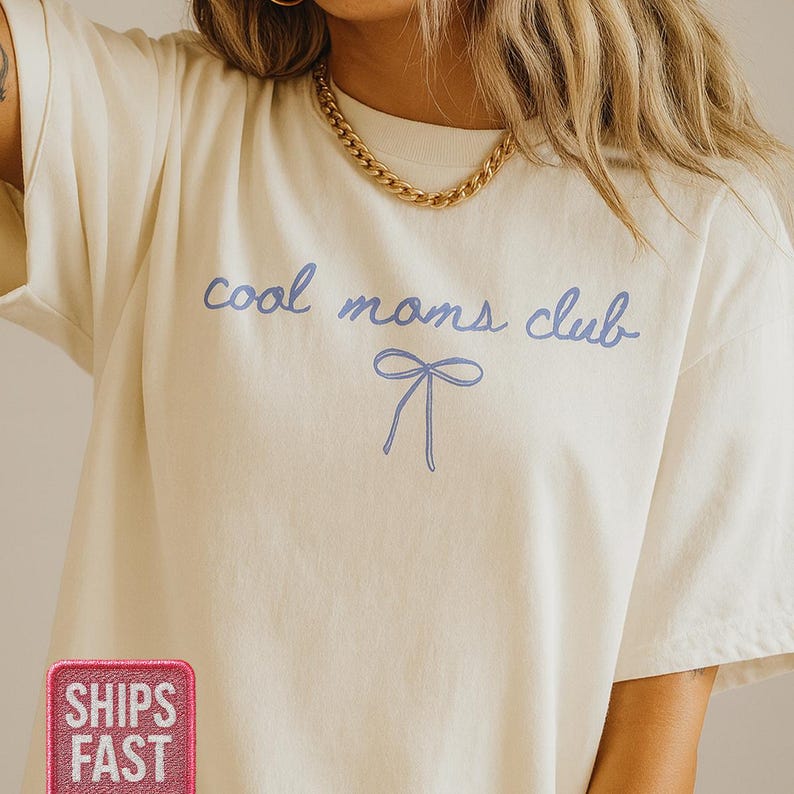 May include: Cream-colored t-shirt with the text "cool moms club" in light blue script, accompanied by a matching bow graphic. The shirt has a relaxed fit and short sleeves, perfect for casual wear.