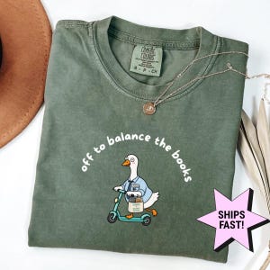 Op de afbeelding: Een olijfgroen t-shirt met een afbeelding van een witte gans in een blauw overhemd met stropdas op een blauwgroene step. De tekst "off to balance the books" staat in witte letters op het shirt. Het shirt heeft een Comfort Colors label in de nek.