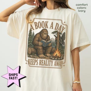 May include: An ivory t-shirt featuring a graphic of a brown Bigfoot sitting on a log reading a book in a forest. A campfire burns nearby, and a moth-like creature is perched on a tree. The text reads A BOOK A DAY KEEPS REALITY AWAY.
