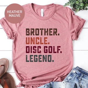 Może przedstawiać: Koszulka w kolorze heather mauve z napisem "Brother. Uncle. Disc Golf. Legend." w stylu vintage.
