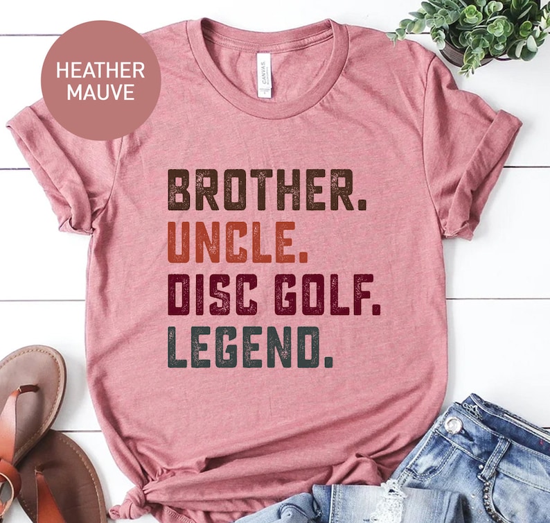 Może przedstawiać: Koszulka w kolorze heather mauve z napisem "Brother. Uncle. Disc Golf. Legend." w stylu vintage.