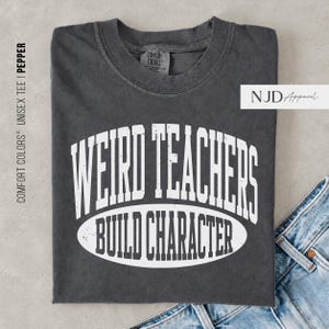 以下が含まれることがあります： クルーネックのチャコールグレーの半袖Tシャツ。前面には、楕円形の枠の中に太字のアーチ状フォントで「WEIRD TEACHERS BUILD CHARACTER」と書かれた、ヴィンテージ風の白いテキストがプリントされています。全体的に色あせた風合いの仕上げです。