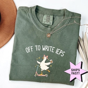 Könnte beinhalten: Olivgrünes T-Shirt mit dem Text "OFF TO WRITE IEPs" und einer Cartoon-Ente auf einem Roller, die eine Kaffeetasse hält. Das Shirt hat einen Rundhalsausschnitt und kurze Ärmel. Eine goldene Halskette liegt über dem Shirt.