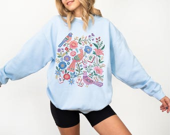 May include: A light blue crewneck sweatshirt featuring a colorful graphic print of three birds, various flowers, and several mahjong tiles with Chinese characters. The design includes pink, red, blue, and green floral elements.
