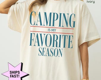 May include: Ivory-colored t-shirt with the text "CAMPING IS MY FAVORITE SEASON" in blue. The text is separated by a red line. The words "comfort colors: ivory" are in the upper right corner. A star-shaped graphic with the words "SHIPS FAST!" is in the lower left corner.