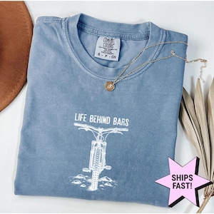 Op de afbeelding: Een blauw Comfort Colors T-shirt met de tekst "LIFE BEHIND BARS" en een witte mountainbike-afbeelding. Een gouden ketting met een hanger ligt op het shirt. Het shirt is opgevouwen en heeft een "SHIPS FAST!" sticker.