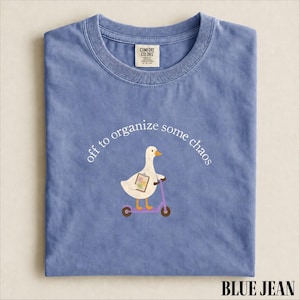 以下が含まれることがあります： 青いジーンズ色のTシャツで、クリップボードを持って紫色のスクーターに乗っている白いガチョウのグラフィックが描かれています。ガチョウの上のテキストには、off to organize some chaosと書かれています。シャツの首元にはComfort Colorsのブランドラベルが付いています。