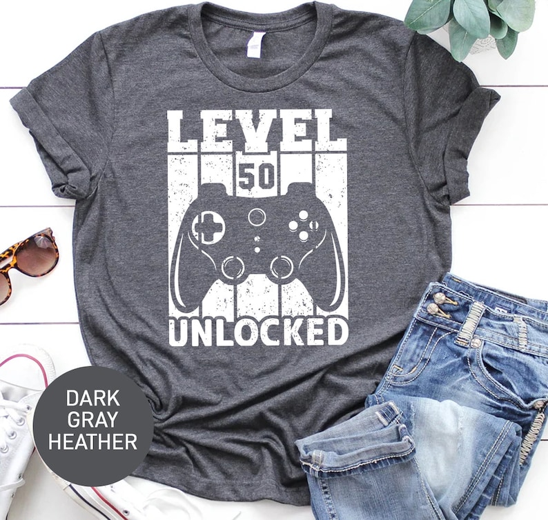 May include: Dark gray heather t-shirt with a white graphic of a video game controller and the text "Level 50 Unlocked".
