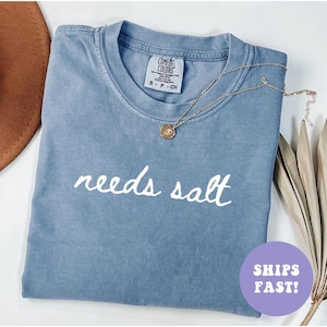 May include: A blue Comfort Colors t-shirt with the words "needs salt" in white script. A gold necklace with a pendant is on the shirt. The shirt is folded and displayed with a brown hat and dried plant.