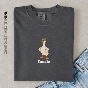 May include: A charcoal gray crew neck t-shirt featuring a graphic of a white duck wearing a green baseball cap and black sunglasses. Below the duck, the word funcle is printed in white lowercase letters. The shirt has a Comfort Colors label.
