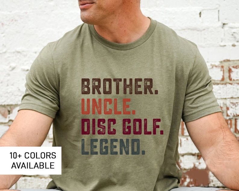 Może przedstawiać: Zielony T-shirt z tekstem "Brother. Uncle. Disc Golf. Legend." w stylu vintage. Tekst jest w odcieniach brązu, pomarańczy, czerwieni i szarości.