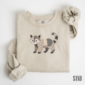 Può includere: Una felpa girocollo color sabbia con un disegno di gatto ricamato. Il gatto è composto da vari inserti in tessuto nei toni del marrone, beige e nero, inclusi motivi a quadretti, floreali e a tinta unita, con una lunga coda ricurva.
