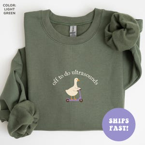 May include: A light green crewneck sweatshirt featuring an embroidered graphic of a white goose riding a purple scooter. The text "off to do ultrasounds" is arched above the goose in white lettering. The garment has long sleeves and a ribbed collar.