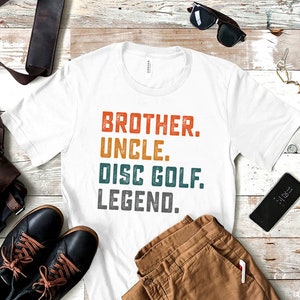 Może przedstawiać: Biała koszulka z nadrukiem w stylu vintage, kt&oacute;ry głosi "Brother. Uncle. Disc Golf. Legend."