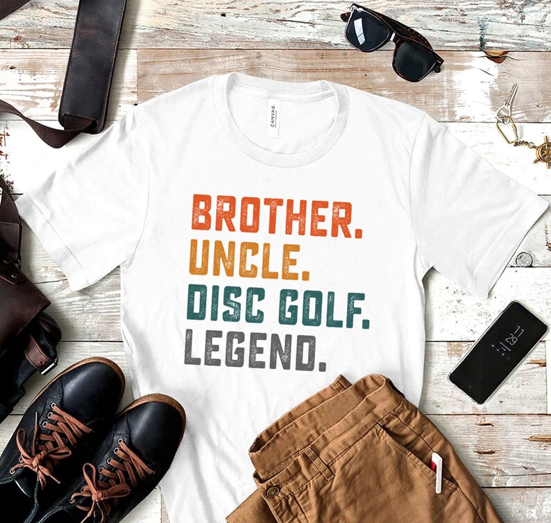 Może przedstawiać: Biała koszulka z nadrukiem w stylu vintage, kt&oacute;ry głosi "Brother. Uncle. Disc Golf. Legend."