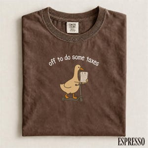 Może przedstawiać: Brązowy t-shirt z okrągłym dekoltem i grafiką przedstawiającą jasnobrązową kaczkę na szarej hulajnodze, trzymającą dokument z napisem ITR. Tekst nad kaczką brzmi off to do some taxes białymi literami. Koszulka posiada metkę marki Comfort Colors.