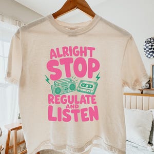 Può includere: Una t-shirt boxy color avorio con una stampa grafica che riporta il testo ALRIGHT STOP REGULATE AND LISTEN in rosa, accompagnato da un'illustrazione verde di un boombox e di una cassetta. La maglietta presenta un girocollo e maniche corte.