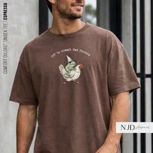 May include: A brown short-sleeve t-shirt featuring a graphic of a green frog wearing a pointed hat while riding a white chicken. The text above the illustration reads, Off to commit dad foolery, in a simple white font.