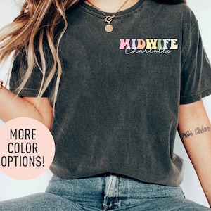 May include: A dark grey t-shirt with the text "MIDWIFE Charlotte" in rainbow colours. The shirt is worn by a person with long blonde hair and a necklace. The text "MORE COLOR OPTIONS!" is in a circle on the left side of the image.