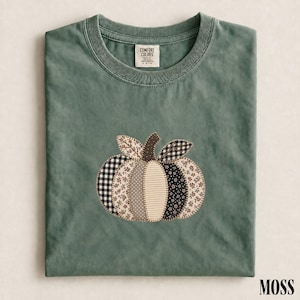 May include: A moss green crew neck t-shirt featuring a central pumpkin graphic composed of various fabric patches. The pumpkin design includes black and white gingham, floral, and striped patterns with visible stitching along the edges.