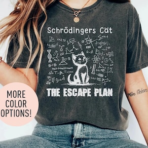 Könnte beinhalten: Ein dunkelgraues T-Shirt mit einer weißen Grafik eines Katers und wissenschaftlichen Formeln. Der Text "Schrödingers Cat" und "The Escape Plan" ist auf dem T-Shirt aufgedruckt.