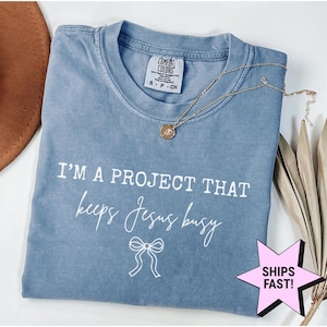 May include: A blue Comfort Colors t-shirt with the text "I'M A PROJECT THAT keeps Jesus busy" in white script. A gold necklace with a pendant rests on the shirt. A pink star-shaped graphic with the words "SHIPS FAST!" is visible.