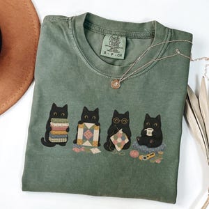Può includere: Una t-shirt verde oliva con una grafica di quattro gatti neri impegnati in attività di quilting, tra cui tenere pile di tessuti, un blocco di trapunta, lavorare a maglia e bere da una tazza. L'etichetta interna sul collo riporta Comfort Colors S-P-CH.