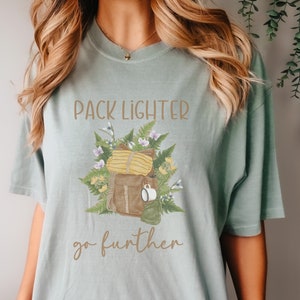 May include: A light blue t-shirt with a brown and yellow illustration of a backpack, flowers, and leaves. The text "PACK LIGHTER go further" is printed on the shirt.