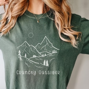May include: A green t-shirt with a white mountain range graphic and the text "Crunchy Outsider".