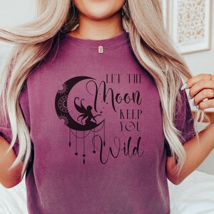May include: A purple t-shirt featuring a black graphic of a crescent moon with a fairy inside. The text "Let the Moon Keep You Wild" is printed on the shirt.