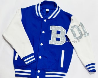 Può includere: Una giacca da baseball blu e bianca con lettere in glitter argentate che dicono "B 01".
