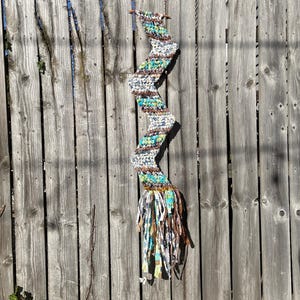 Op de afbeelding: Een handgemaakte, zigzagvormige wandhanger. Het textielkunstwerk is gemaakt van dik garen in de kleuren wit, bruin, blauwgroen en groen. De onderkant heeft een franje van stoffen stroken. De wandhanger wordt tentoongesteld tegen een verweerde houten schutting.