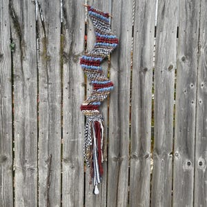 Op de afbeelding: Een handgemaakte textiele wandhanger in een zigzagpatroon. Het stuk heeft een combinatie van bordeauxrode, blauwe, bruine en crèmekleurige garens. De onderkant heeft een kwast van stoffen stroken in verschillende patronen en kleuren. Twee houten breinaalden zitten bovenaan.