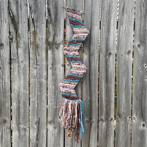 Op de afbeelding: Een handgemaakte, zigzagvormige wandhanger. Het textielkunstwerk heeft een combinatie van bruin, blauw en roze garen. De onderkant van het stuk heeft franjes in vergelijkbare kleuren. De wandhanger wordt tentoongesteld tegen een verweerde houten schutting.