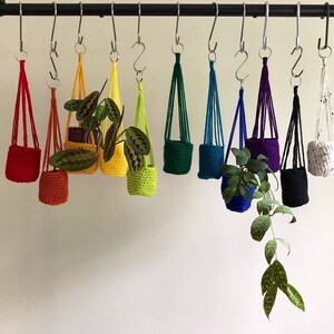 May include: A rainbow assortment of 10 hanging macrame plant hangers with crocheted pots. The hangers are made with different colored cords and the pots are in various colors, including red, orange, yellow, green, blue, purple, and black.