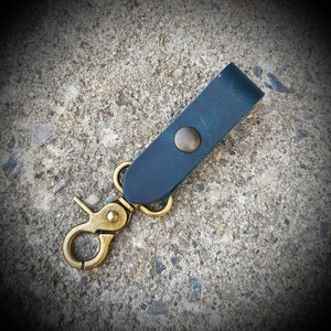 May include: A dark blue leather key chain with a brass hook and a brass ring. The key chain is on a gray concrete surface.