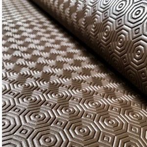 Könnte beinhalten: Eine Rolle brauner, strukturierter Regalboden mit einem sich wiederholenden geometrischen Muster aus Sechsecken, Kreisen und Quadraten. Das Muster ist geprägt und erzeugt eine taktile Oberfläche. Der Liner ist teilweise gerollt.