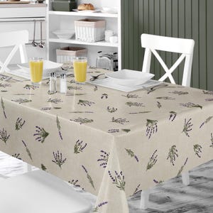 Pode incluir: Uma toalha de mesa retangular bege com um padrão de lavanda. A toalha de mesa está colocada numa mesa branca com cadeiras brancas. A imagem também mostra copos de sumo de laranja, saleiros e pimenteiros e pratos brancos.