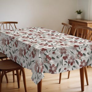 Peut inclure: Nappe rectangulaire à motifs floraux. Le motif présente des fleurs bordeaux, grises et blanches avec des feuilles vertes sur un fond gris clair. La nappe est posée sur une table en bois avec des chaises en bois.