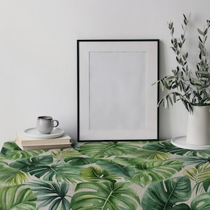 May include: A table with a green and white tropical leaf print tablecloth. A framed picture, a small cup and saucer, a stack of books, and a white jug with greenery are on the table.