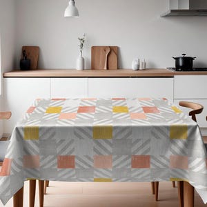 Op de afbeelding: Een grijs tafelkleed met een geometrisch patroon van roze, gele en grijze vierkanten. Het tafelkleed ligt op een houten tafel met twee stoelen.