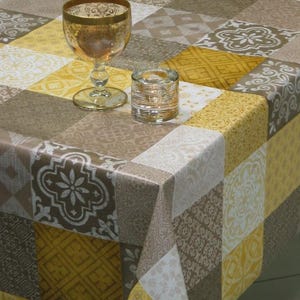 Puede incluir: Un mantel decorativo con un diseño de patchwork en tonos amarillos, marrones y blancos. El mantel presenta una variedad de patrones geométricos y florales. Una copa de vino con borde dorado y un pequeño portavelas de cristal están sobre la mesa.