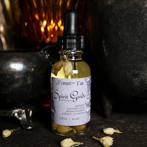 May include: A clear glass bottle with a black dropper top and a purple label that reads "of Forest and Fae Spirit Guide anointing oil guidance communication deepen intuition enhance connection 1 fl oz | 30 ml". The bottle is filled with a light yellow liquid and contains small white flowers.