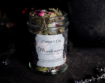 May include: A glass jar filled with dried herbs and flowers, labeled "Manifestation spellwork herb blend" by "of Forest and Fae". The label also includes the text "To focus intention, amplify energy work, visualize desires."