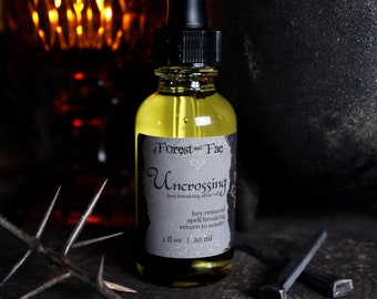 Può includere: Una piccola bottiglia di vetro con un contagocce, riempita di un liquido giallo. La bottiglia ha un'etichetta nera con testo bianco che dice "of Forest and Fae Uncrossing hex breaking altar oil hex removal spell breaking return to sender 1 fl oz | 30 ml".