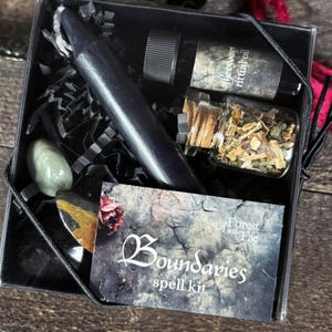 K&ouml;nnte beinhalten: Eine schwarze Box mit einer schwarzen Kerze, einer kleinen &Ouml;lflasche, einem kleinen Glas mit Kr&auml;utern und einer Karte mit der Aufschrift "Boundaries spell kit" mit den Worten "Forest and Fae". Die Box enth&auml;lt auch einen gr&uuml;nen Stein und einen schwarzen Stein.