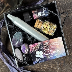 May include: A black box with a silver candle, a small bottle of oil, a glass jar with dried herbs, and three crystals. The box has a label that says "Clarity spell kit" and "Forest of and Ease".