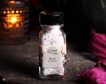 May include: A clear glass bottle with a black screw-top lid containing white salt mixed with small dried pink rose petals. The label features the text Forest of and Fae, Rose, and Ritual Salt, with a decorative floral border design.