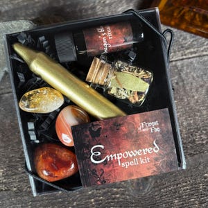 May include: A black box with a gold candle, three red carnelian stones, a clear glass jar with herbs, and a small bottle of oil. The box has a red and black label that says "Empowered spell kit" and "of Forest and Fae".